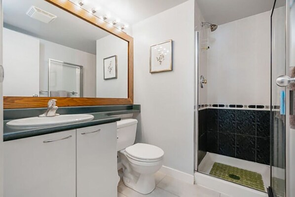 A bathroom with a white sink, toilet, and shower. There is a mirror with lights above it and framed pictures on the wall.