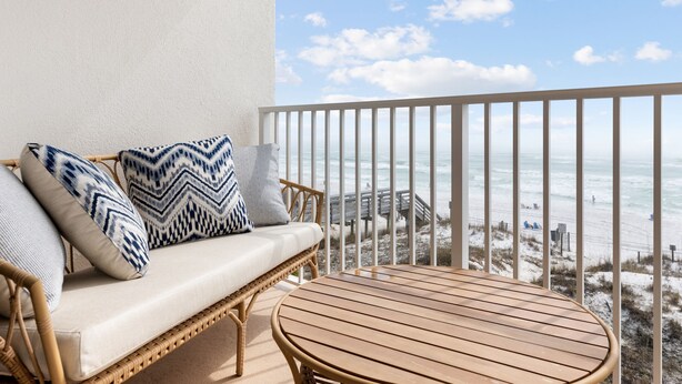 A balcony with a wicker sofa and a wooden table, overlooking a beach with a wooden walkway and beachgoers. - Oceanfront Oasis. Expansive balcony, cozy decor, stunning views over the dunes. (Fort Walton Beach)