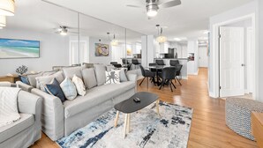A modern living room with a grey sofa, black coffee table, and a blue rug. There is a dining area with a table and chairs, and a kitchen in the background. - Oceanfront Oasis. Expansive balcony, cozy decor, stunning views over the dunes. (Fort Walton Beach)