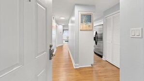 A hallway with white doors, a wooden floor, a refrigerator, and a painting of palm trees. - Oceanfront Oasis. Expansive balcony, cozy decor, stunning views over the dunes. (Fort Walton Beach)