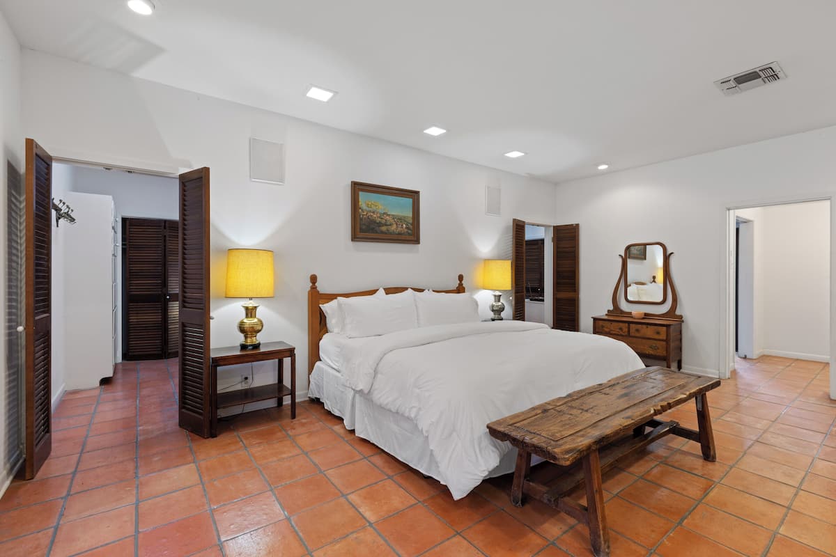 Quarto com cama, mesas de cabeceira, um banco de madeira, um quadro na parede e um espelho.