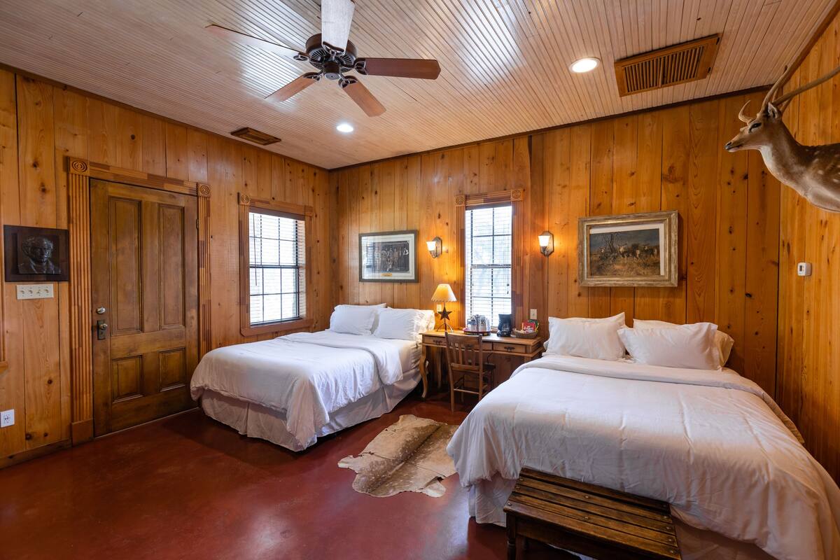 Quarto com paredes de madeira, duas camas, uma escrivaninha, uma cadeira, um ventilador de teto e um chifre de veado fixado na parede.