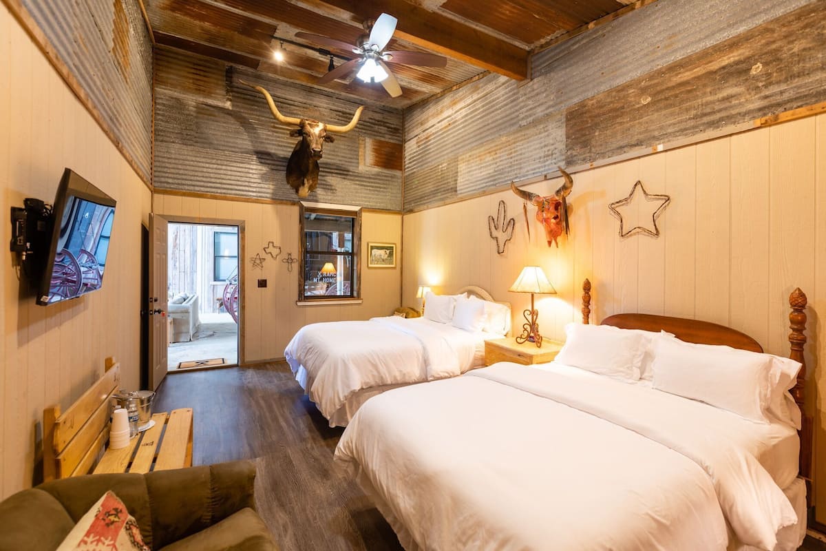 Quarto com duas camas, ventoinha no teto, decoração rústica, televisão e porta que dá para outra área.