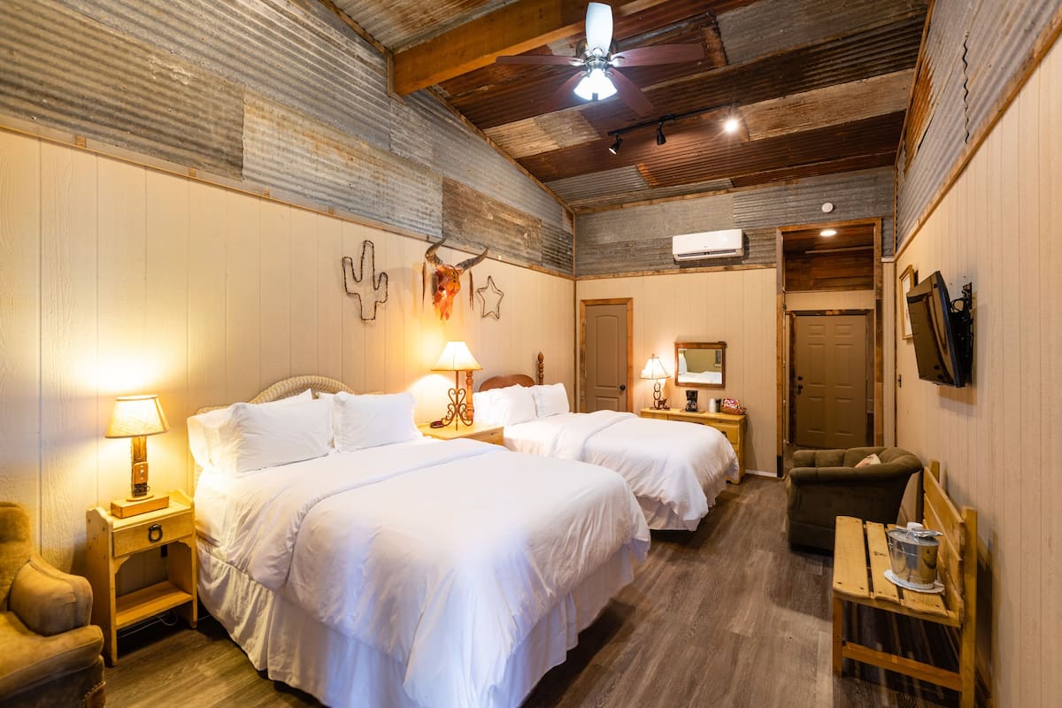 Quarto de hotel com duas camas, um ventilador de teto, uma televisão e uma mesa de cabeceira de madeira.