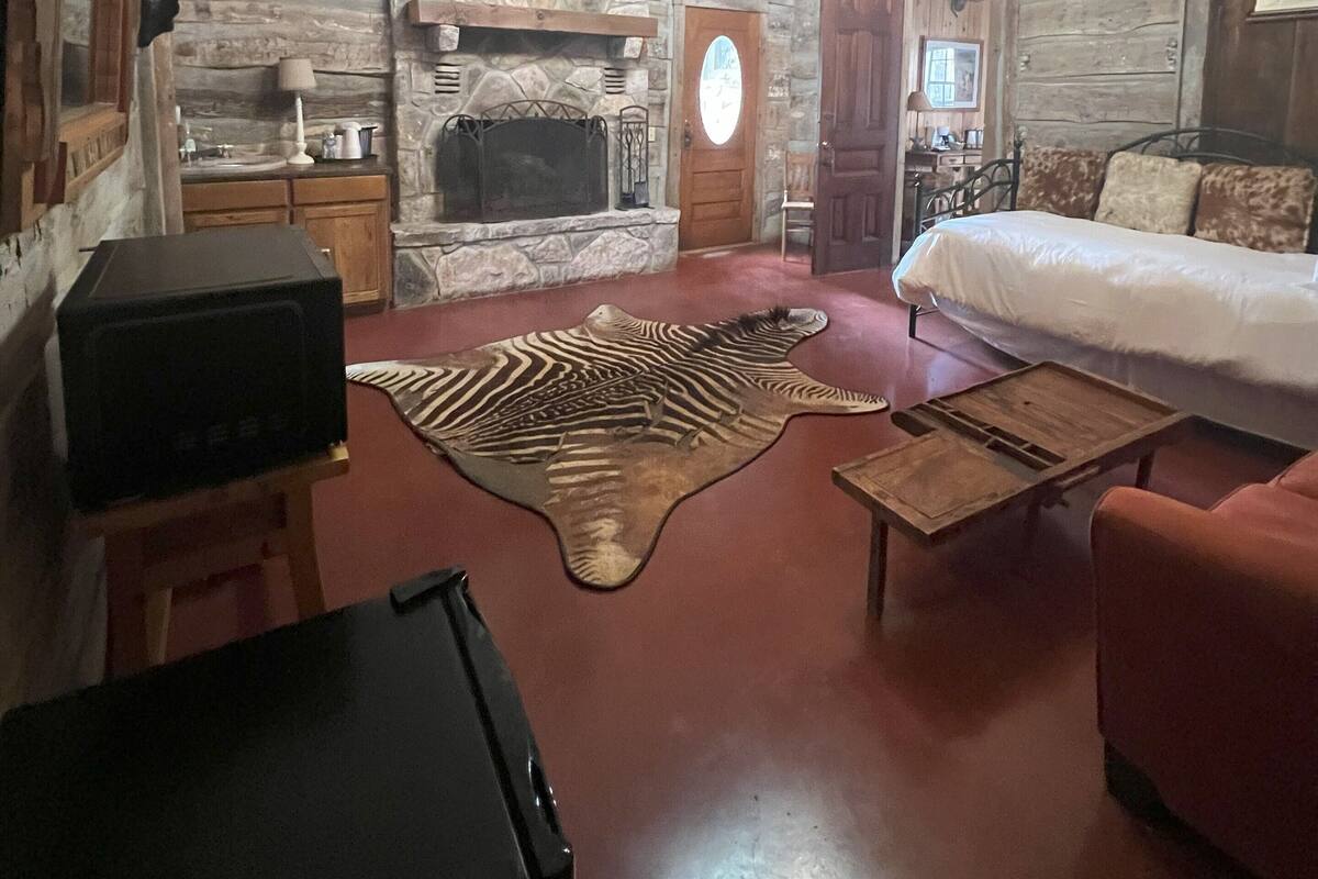 Interior rústico de cabana de madeira com lareira de pedra, cabeça de animal montada, cama, sofá vermelho e televisão sobre suporte.