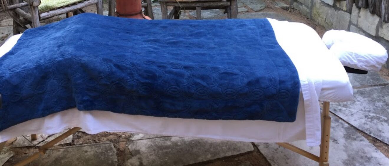 A massage table with a blue blanket and white sheets, set on a stone floor with a wooden frame.