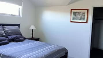 Un dormitorio con cama, ventilador de techo, mesita de noche con lámpara, un cuadro en la pared y una puerta que da a otra habitación.