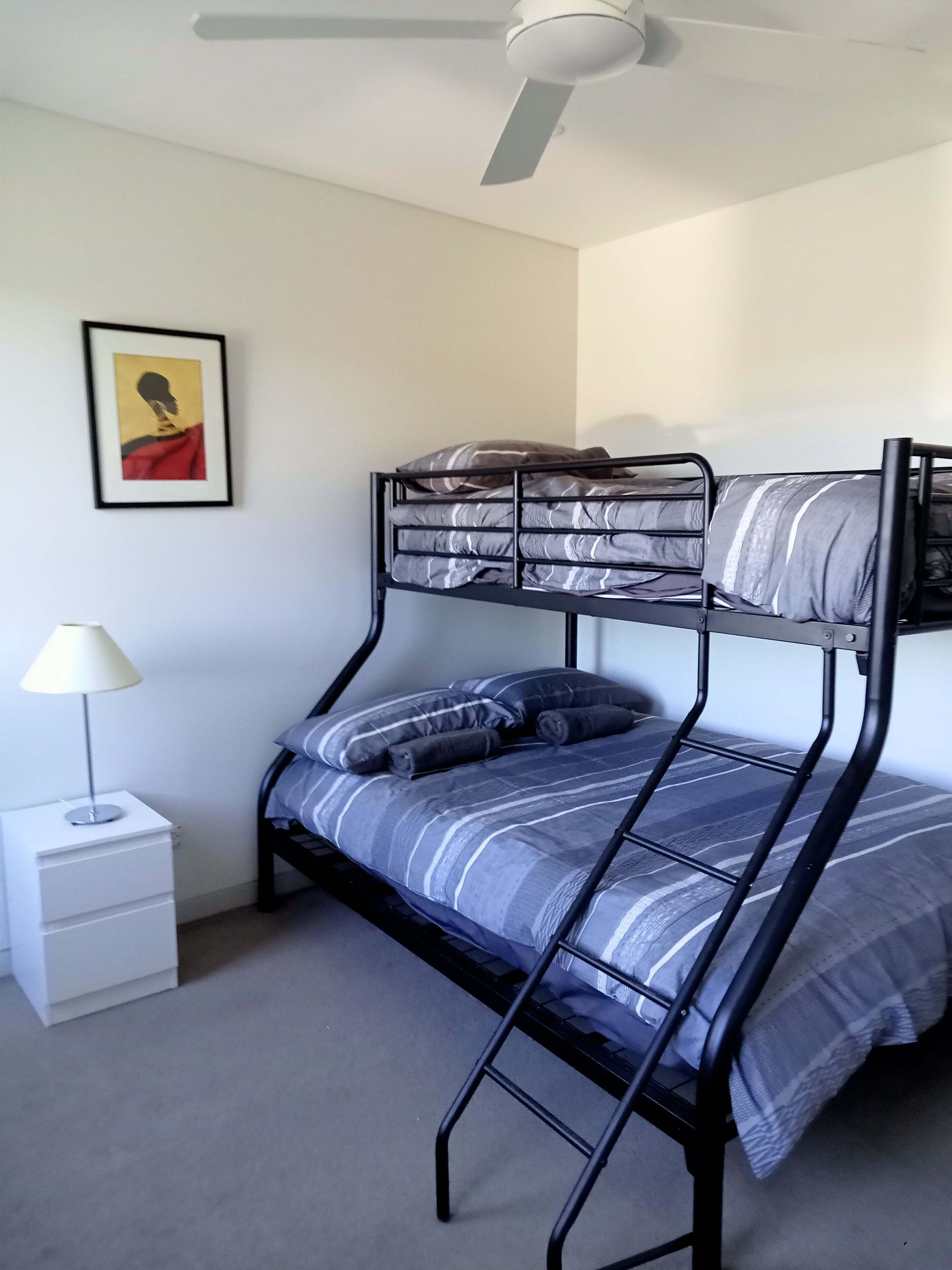 A bunk bed with a ceiling fan, a bedside table with a lamp, and a framed picture on the wall.