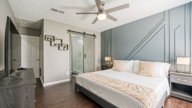 A bedroom with a large bed, a ceiling fan, a nightstand, a dresser, a sliding glass door, and a small plant.