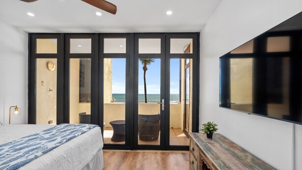 Un dormitorio con cama, cómoda de madera, televisor y puerta corrediza de vidrio que da a un balcón con vista al mar.