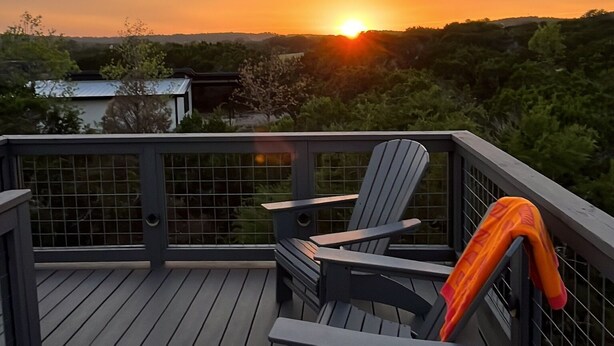 A wooden deck with two Adirondack chairs and a table, overlooking a sunset with the sun near the horizon.