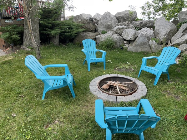 A fire pit with four blue Adirondack chairs arranged around it in a grassy outdoor area with large rocks and trees in the background.