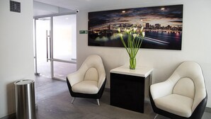 A modern reception area with two white chairs, a black and white desk, a cityscape painting, and a glass door leading outside. - Apartasuite Edificio Monir (Bogotá)
