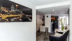 A modern hotel lobby with a cityscape painting, a black leather chair, a small table with a vase of flowers, and a sleek reception desk. - Apartasuite Edificio Monir (Bogotá)