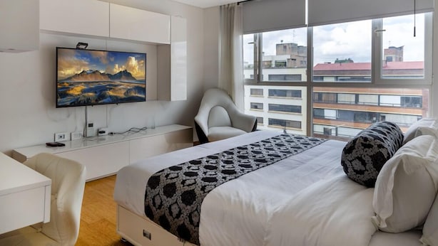 A modern hotel room with a large bed, a TV mounted on the wall, a desk, and a chair. - Apartasuite Edificio Monir (Bogotá)
