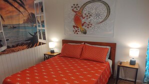 A bedroom with a bed, two bedside tables with lamps, a wall tapestry, and a window with a view of boats and a cliff.