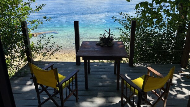 A wooden deck with two yellow chairs and a table, overlooking a clear blue lake with a forested shoreline. - Freshwater Private Resort (Aore Island)