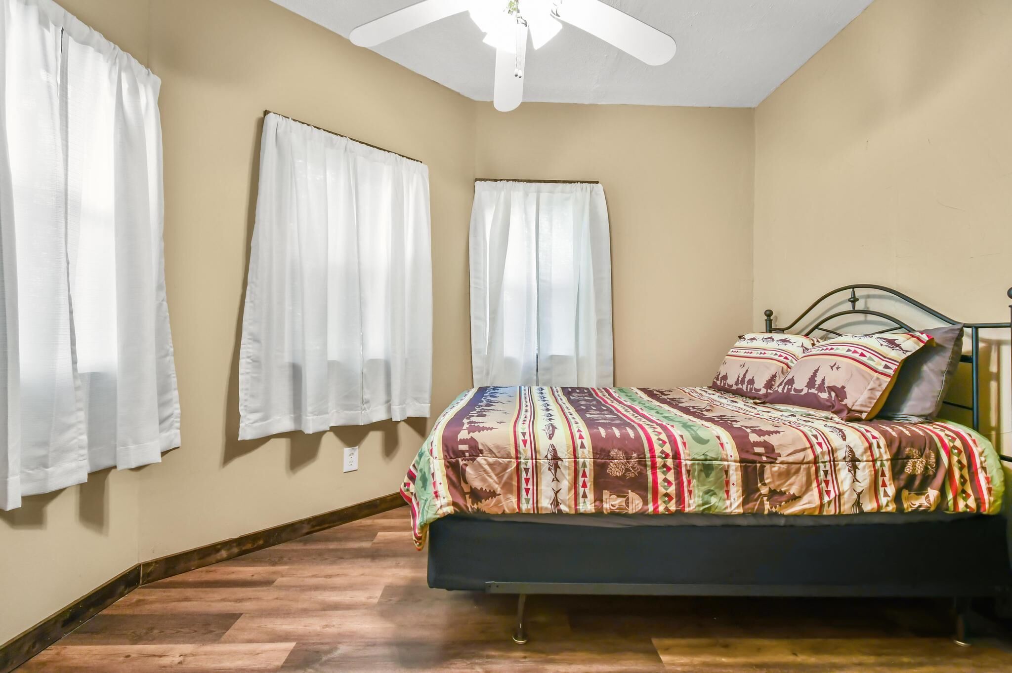 A bedroom with a bed, white curtains, a ceiling fan, and a patterned bedspread.