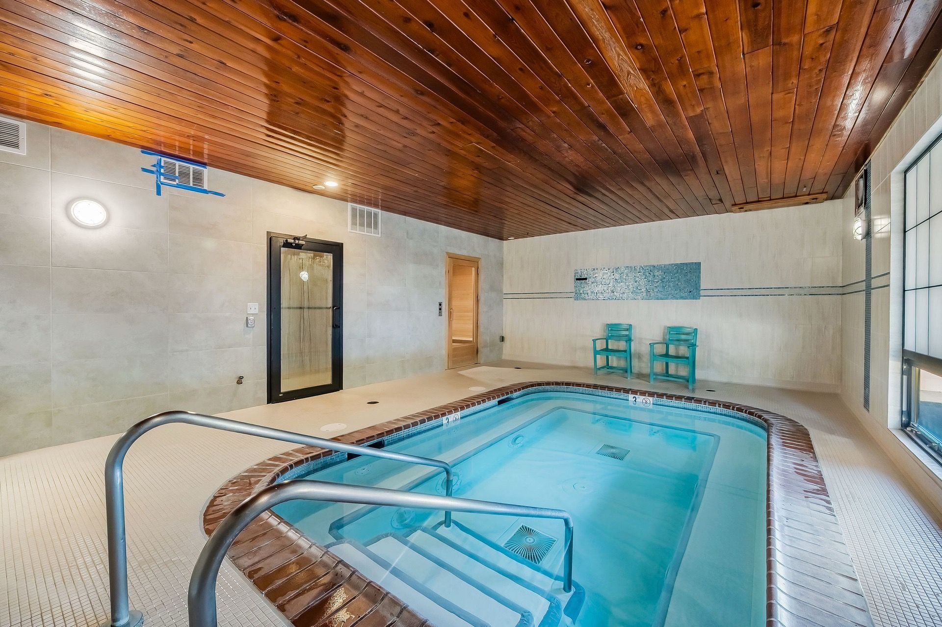 An indoor swimming pool with a wooden ceiling, a glass door, and two turquoise chairs.