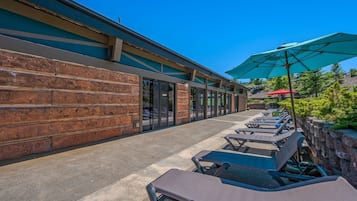 A covered outdoor seating area with lounge chairs and umbrellas, adjacent to a building with a brick facade and large windows.