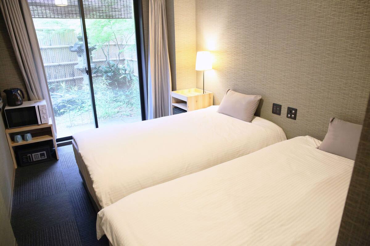 Een hotelkamer met twee bedden, een nachtkastje met lamp, een tafeltje met waterkoker en uitzicht op de omgeving door een schuifdeur.
