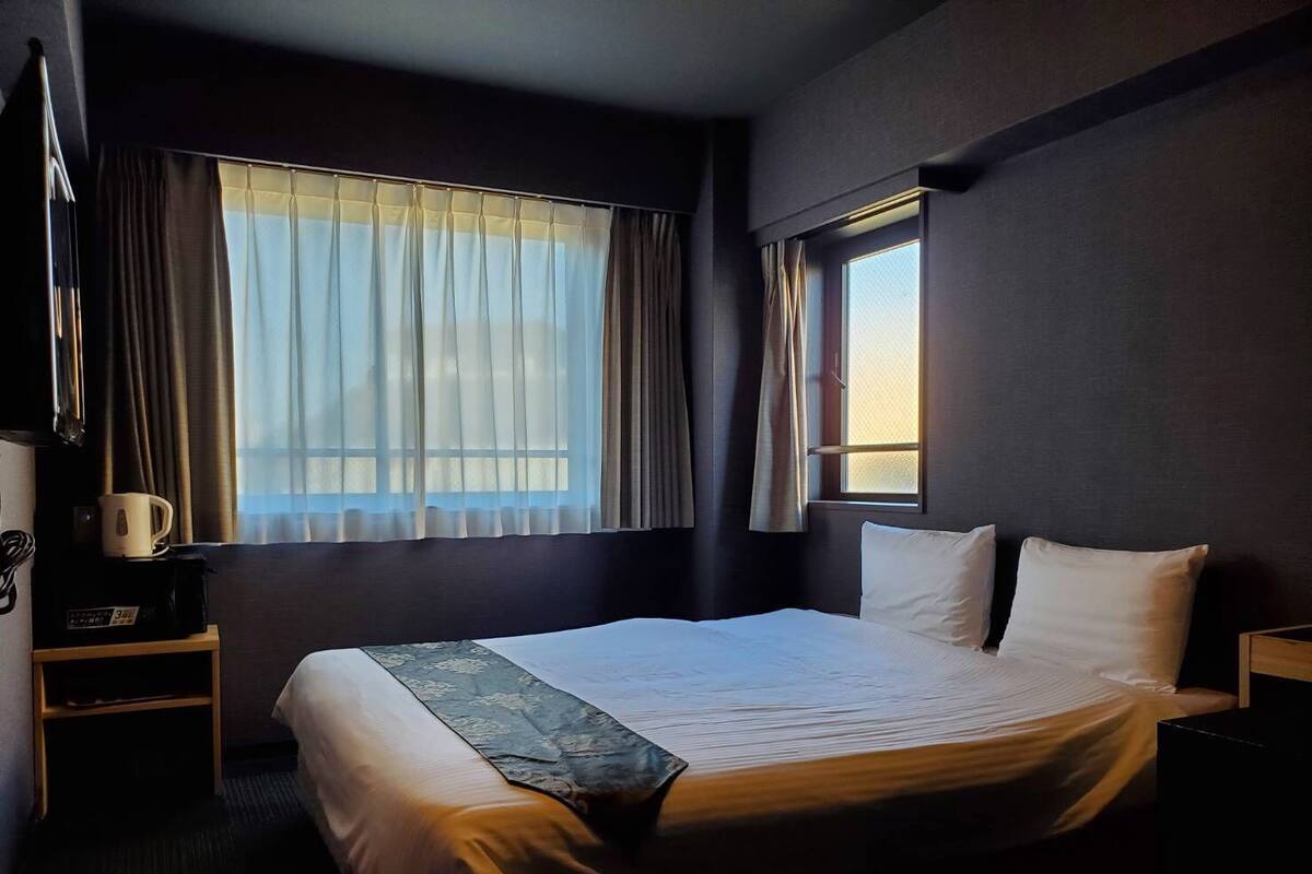 Een hotelkamer met een bed, nachtkastje, koffiezetapparaat en een raam met gordijnen.