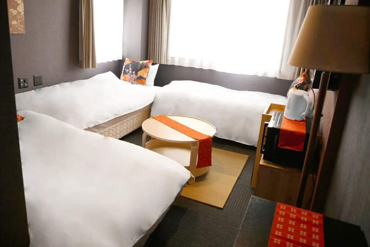 Quarto de hotel com duas camas, uma mesa pequena redonda, uma luminária e uma mesinha de bandeja vermelha.