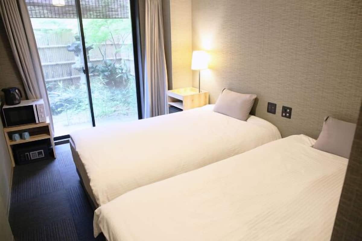 Quarto de hotel com duas camas, uma janela com cortinas, uma mesa de cabeceira com abajur e uma pequena estante com objetos.