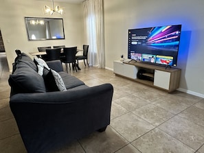 Una sala de estar moderna con un sofá oscuro, un televisor sobre un mueble de madera, una zona de comedor con sillas y un espejo grande en la pared.