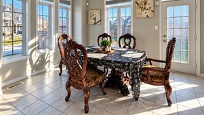 Une salle à manger avec une table dressée pour quatre personnes, entourée de chaises décorées, de grandes fenêtres et offrant une vue sur un quartier résidentiel.