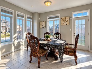 A dining room with a table set for four, surrounded by ornate chairs, large windows, and a view of a residential area.
