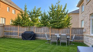 Un jardin avec une clôture en bois, un barbecue couvert et une table à manger avec des chaises.