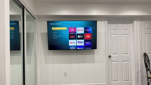 Televisor de pantalla plana montado en una pared blanca que muestra una interfaz de Roku con diversas opciones de transmisión.