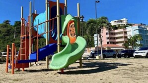 Un'area giochi con scivolo e strutture per arrampicarsi, con sullo sfondo un edificio e auto parcheggiate.