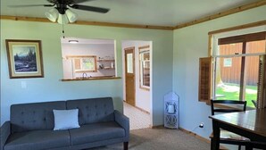A living room with a grey sofa, a dining table with chairs, a ceiling fan, and a view of a backyard through a sliding glass door. - Camas Creek Cabin (Hamilton)