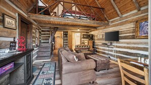 L’intérieur d’une cabane en rondins, avec un escalier en bois, un canapé, une table basse, un téléviseur et une cheminée.