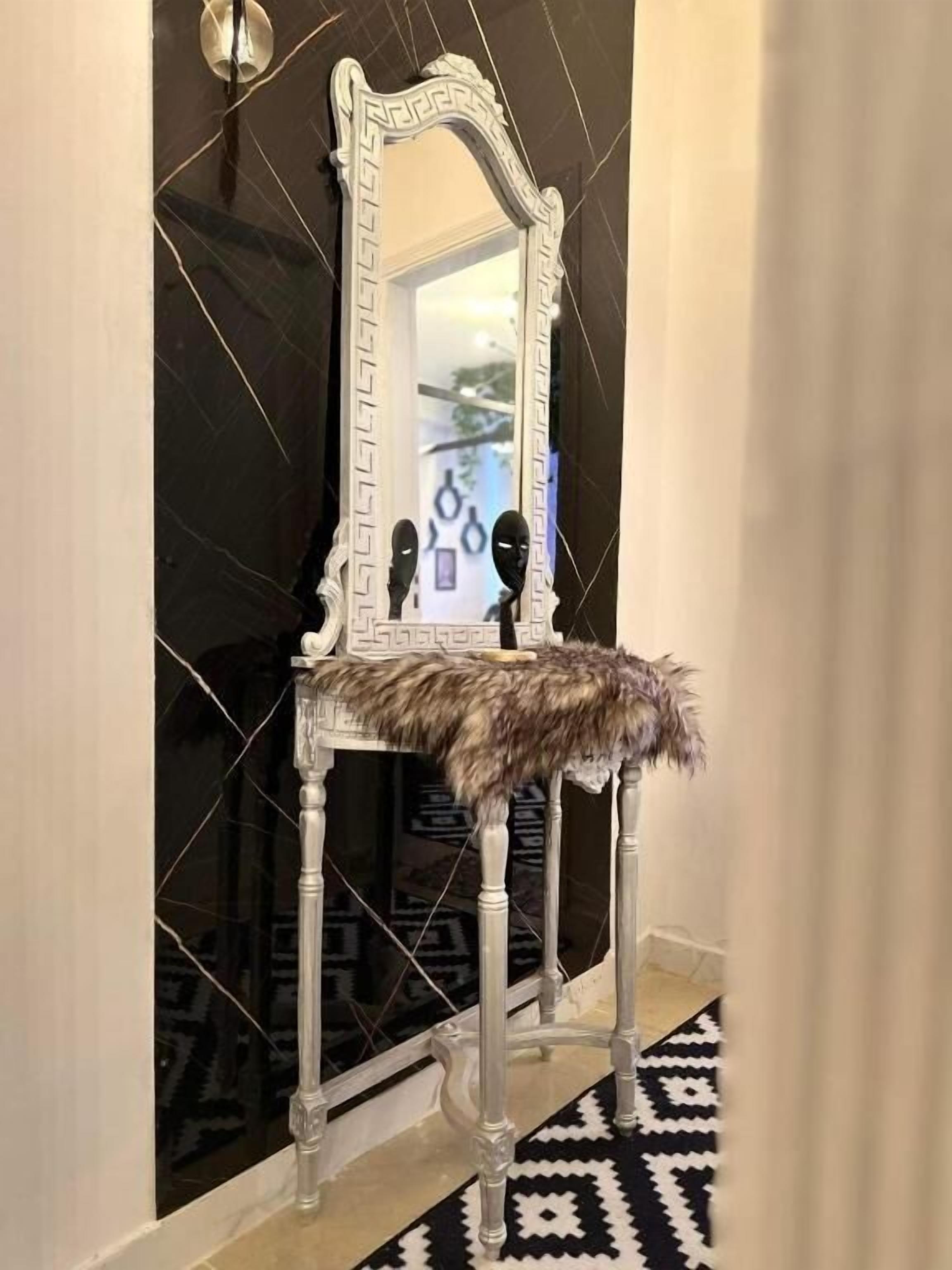 A chair with a fur throw, positioned in front of a mirror with a decorative frame, against a wall with a black and white patterned floor.