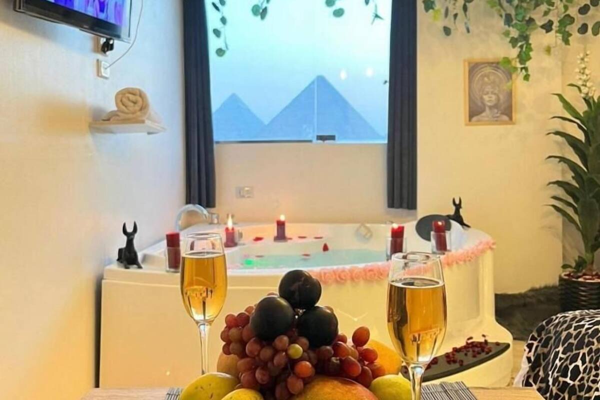 Une chambre avec une baignoire, deux verres de vin, un panier de fruits et un téléviseur fixé au mur.