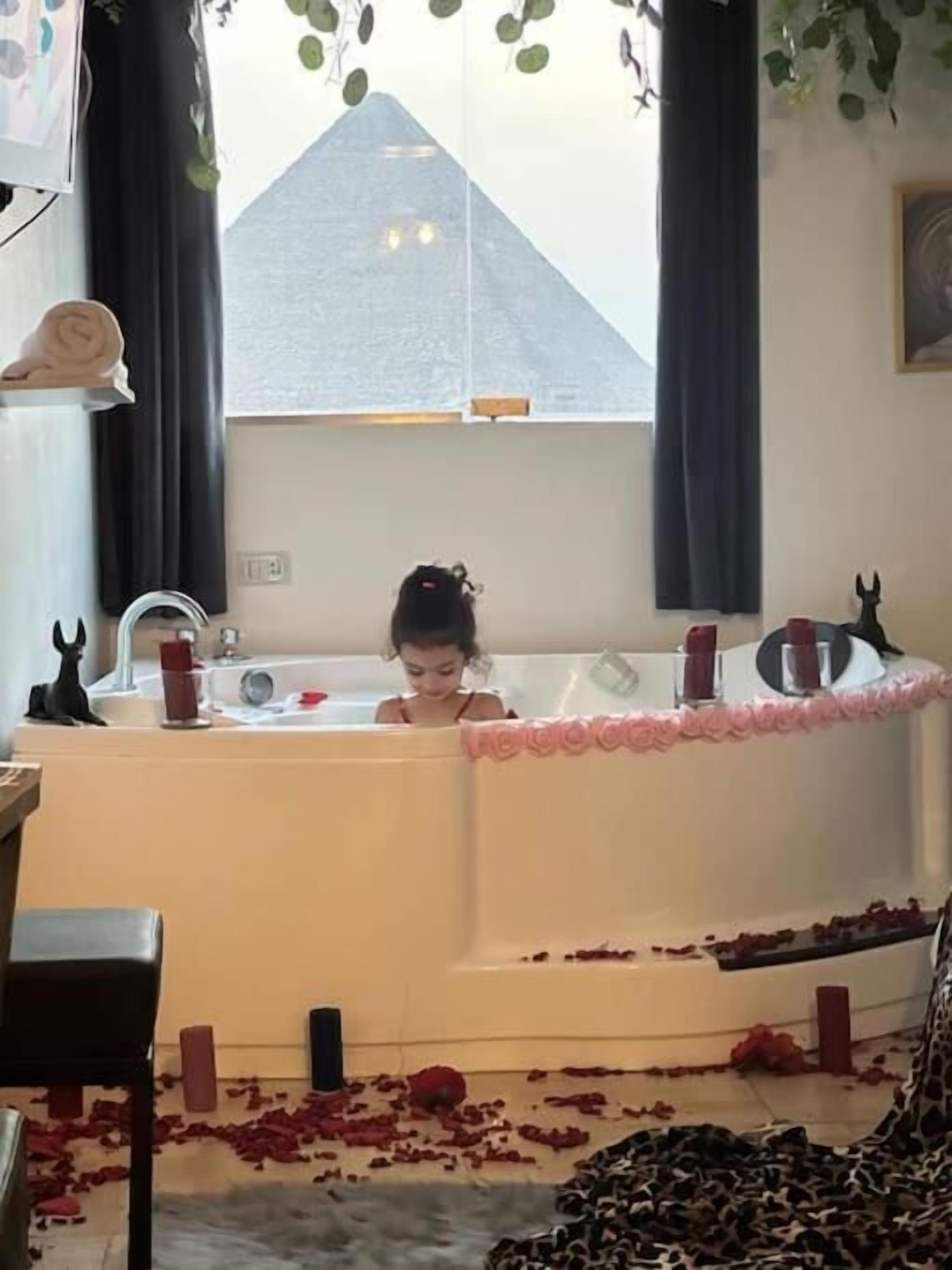 A room with a bathtub filled with rose petals, a window with curtains, and a leopard print rug on the floor.