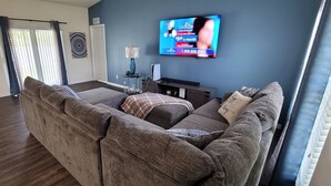 A living room with a sectional sofa, a flat-screen TV mounted on the wall, and a small side table with a lamp.