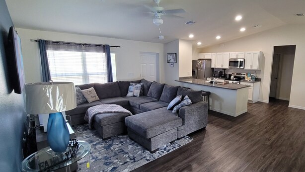 A modern living room with a sectional sofa, a dining area, and a kitchen with white cabinets and stainless steel appliances.