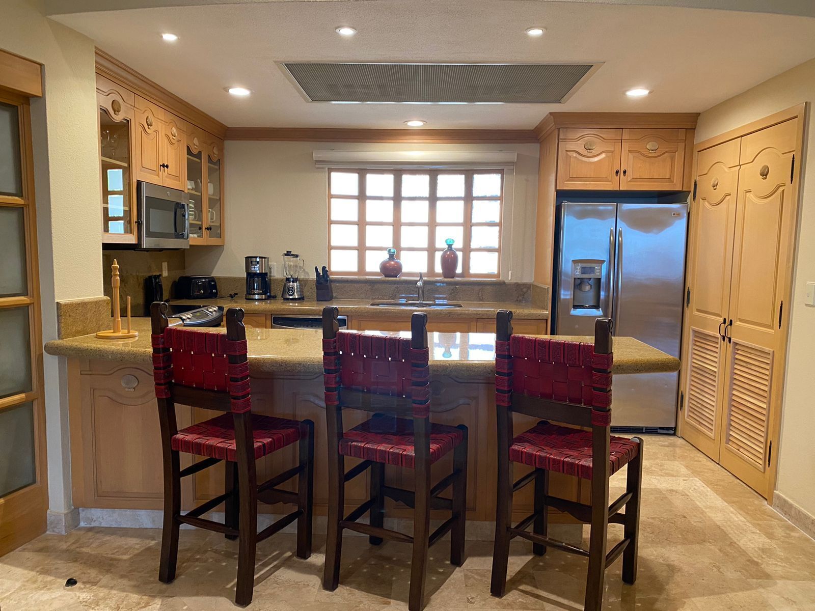 A kitchen with wooden cabinets, a stainless steel refrigerator, a microwave, and a coffee maker. There are three red cushioned chairs at the island.