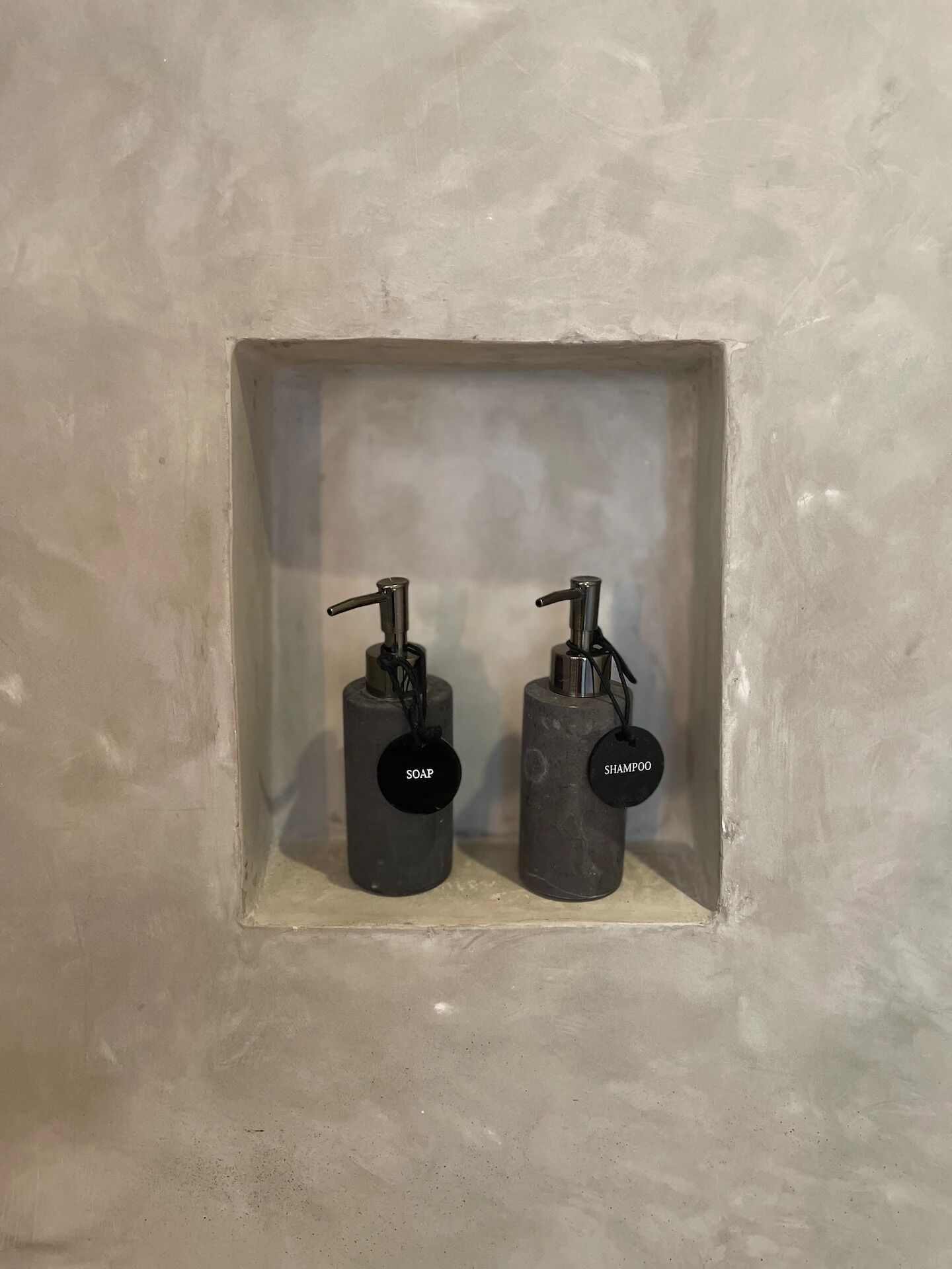 Zwei schwarze Seifenspender mit der Aufschrift 'SOAP' und 'SHAMPOO' in einer rechteckigen, weißen Nische an einer strukturierten Wand.