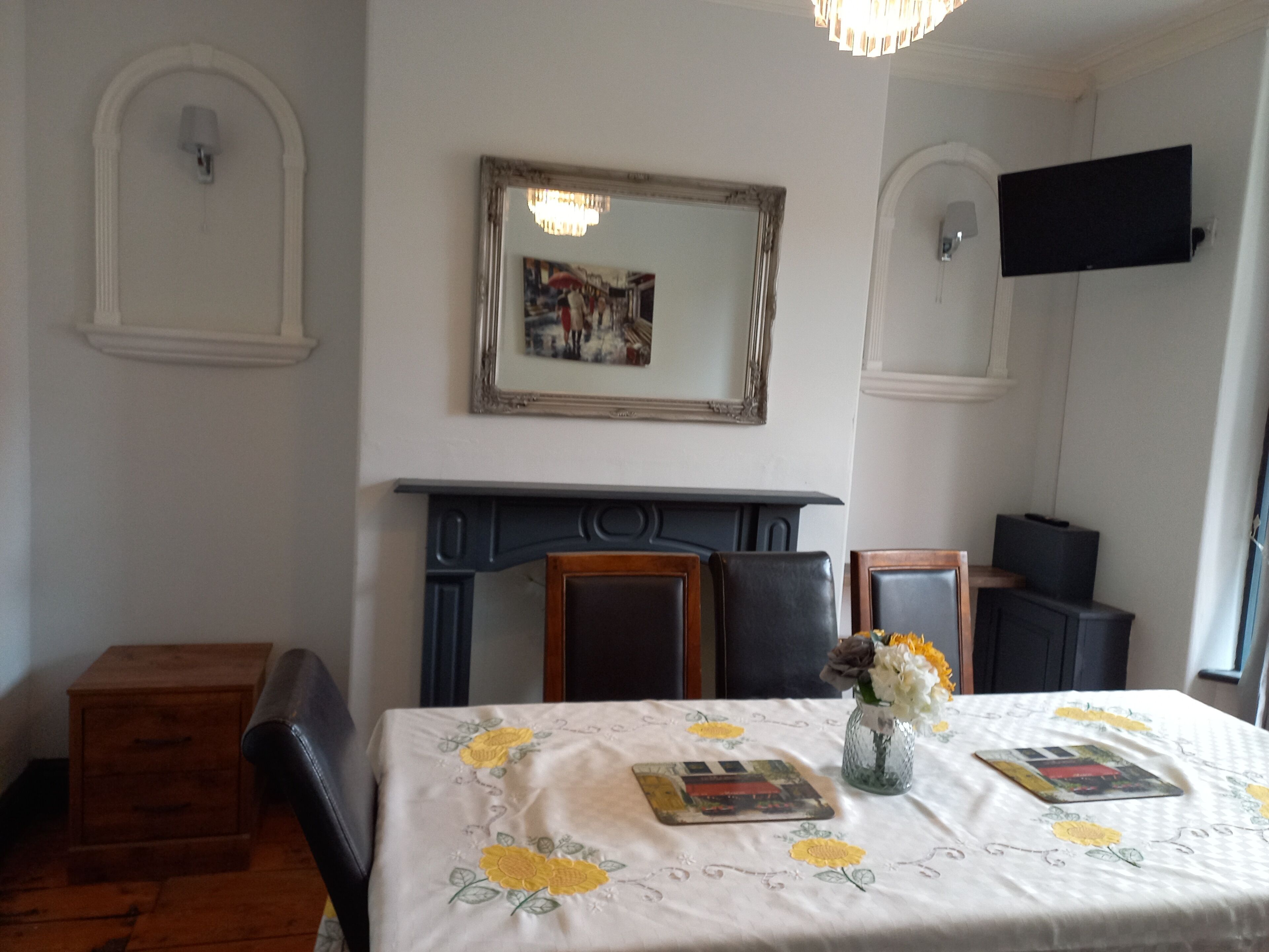 A dining area with a table set for two, a fireplace, a mirror, and a flat-screen TV mounted on the wall.