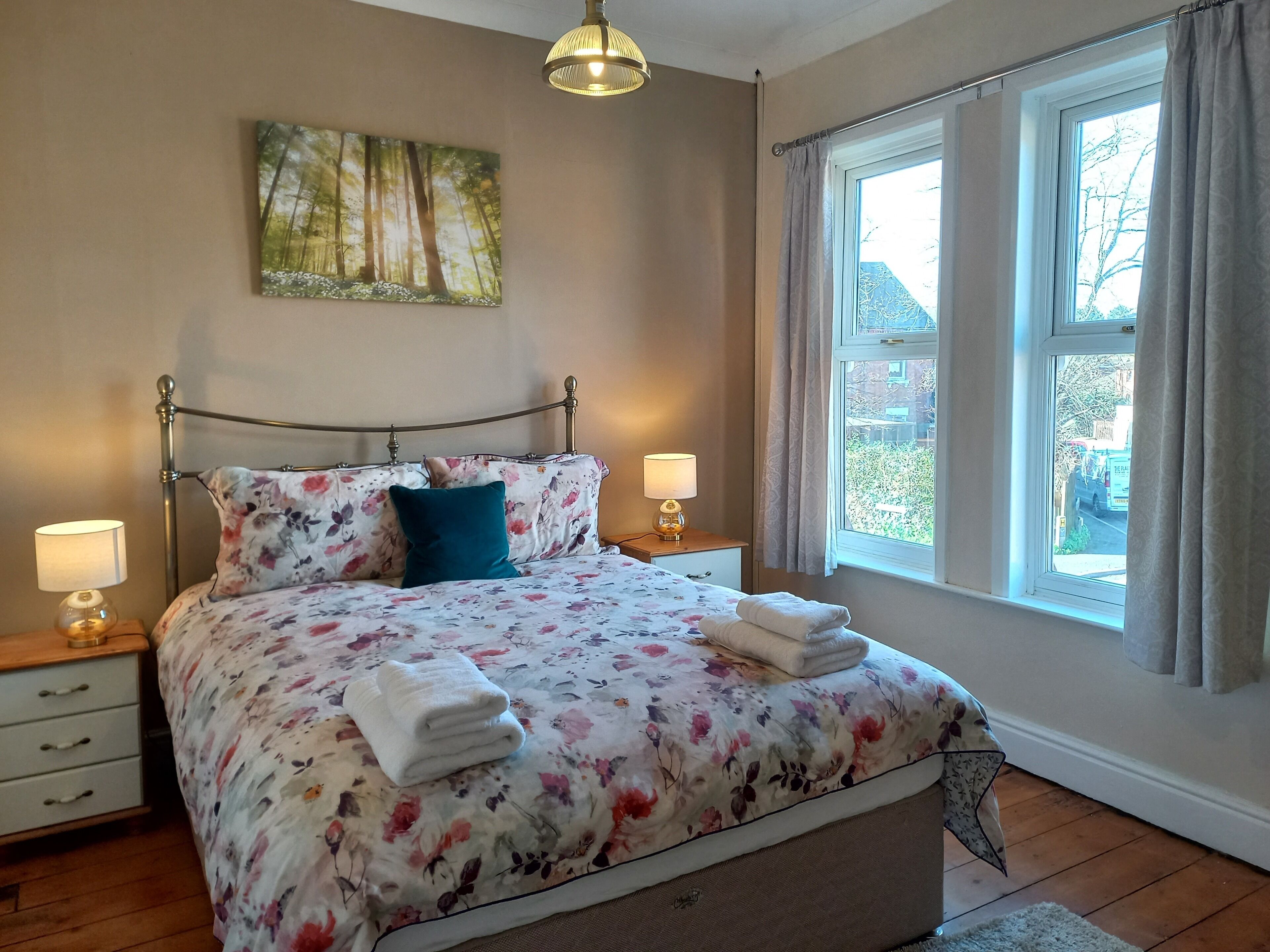 A bedroom with a bed, two bedside tables, a window with curtains, a hanging light fixture, and a painting on the wall.