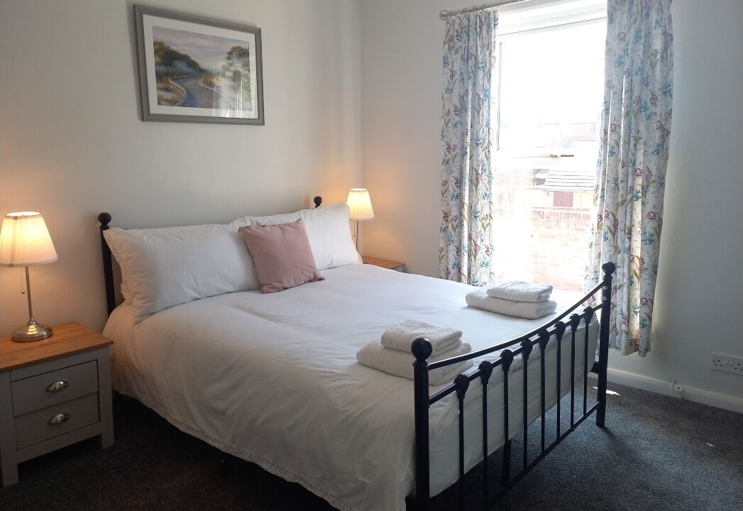 Een opgemaakt bed met witte en roze kussens, een ingelijst beeld aan de muur en een raam met bloemen gordijnen.