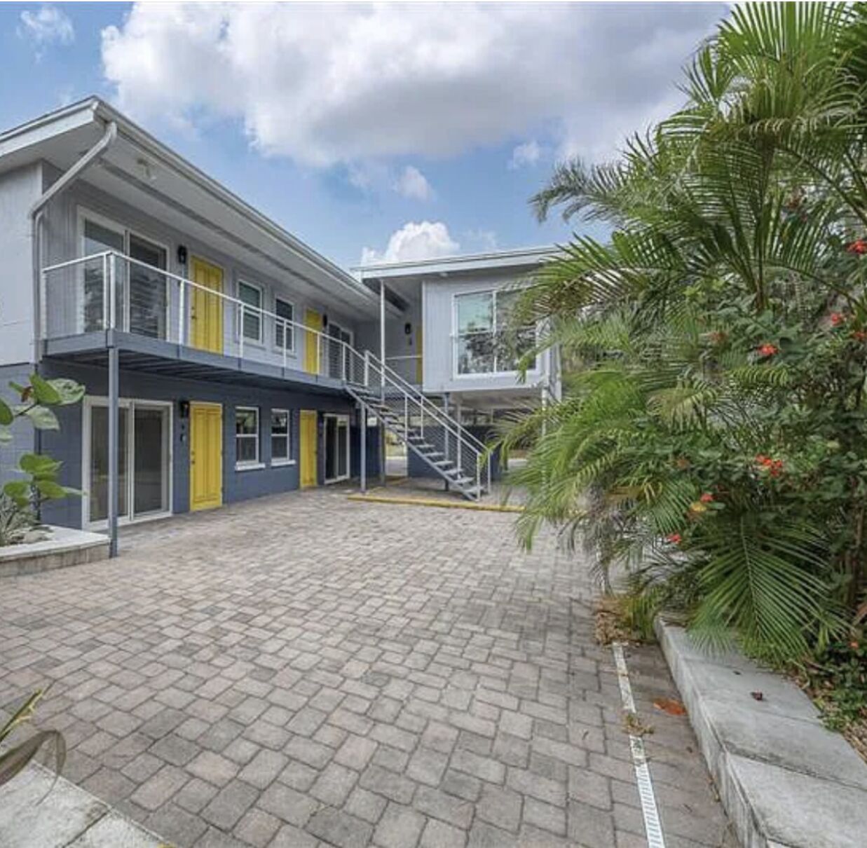 A modern building with a paved courtyard, a staircase, and a small garden area with palm trees.