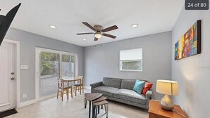 A living room with a grey sofa, a small table, a ceiling fan, and a window.