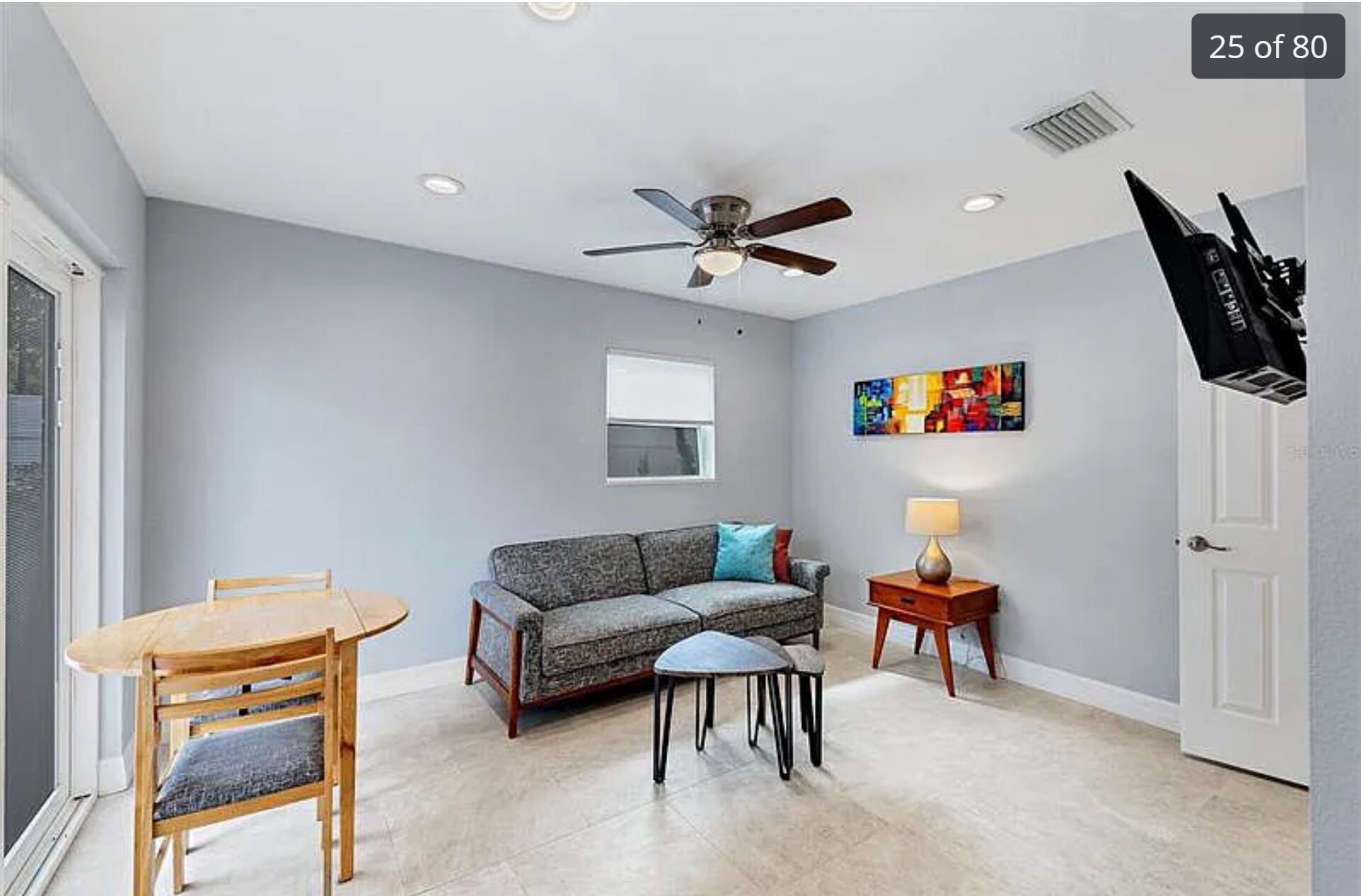 A living room with a ceiling fan, a sofa, a coffee table, a dining table with chairs, a window, and a painting on the wall.