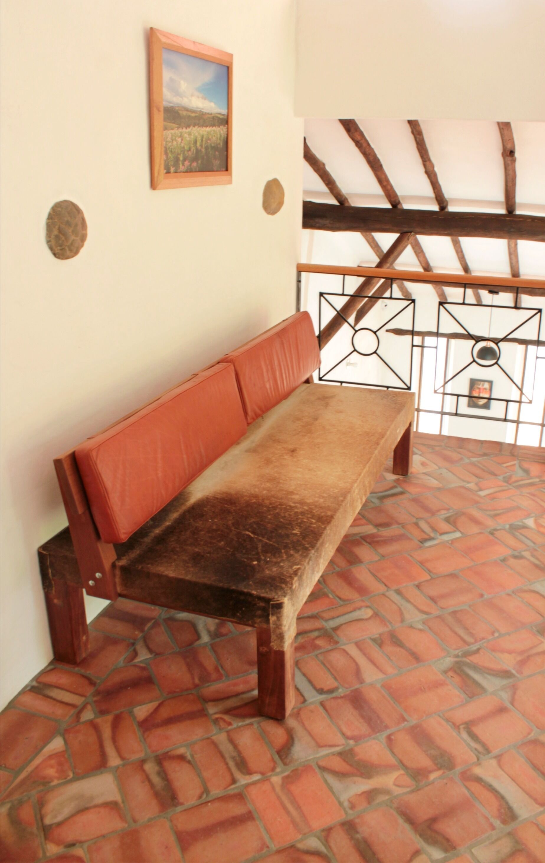 Un banco con un asiento y respaldo desgastados, suelo de baldosas, techo de vigas de madera y un cuadro enmarcado en la pared.